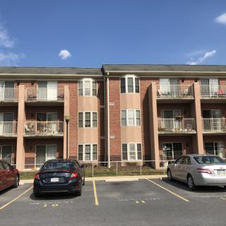 A three-story brick apartment building with balconies and parked cars in front under a clear sky.