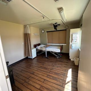 A view of a sparsely furnished medical room with a bed, clock, and refrigerator.