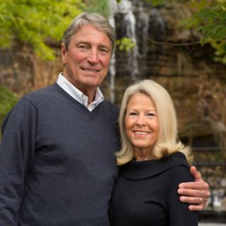 An older couple smiling in front of a waterfall in a natural setting.