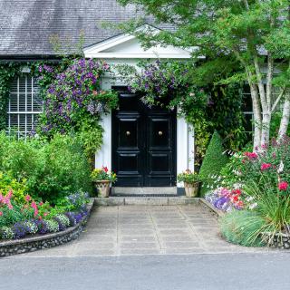 A charming garden path leads to a house with a black double door, surrounded by colorful flowers and lush greenery.