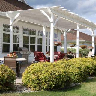 A sunny patio with a white pergola and outdoor seating area, surrounded by well-maintained bushes.