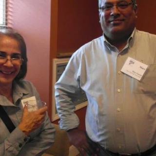 Two individuals at an indoor event wearing name tags, one holding a drink and both smiling.