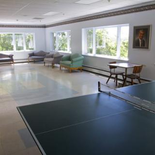 A bright recreation room featuring a ping pong table, sofas, and chairs with large windows letting in natural light.