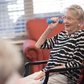 An elderly woman exercises with a dumbbell while seated indoors, smiling and wearing a striped shirt.