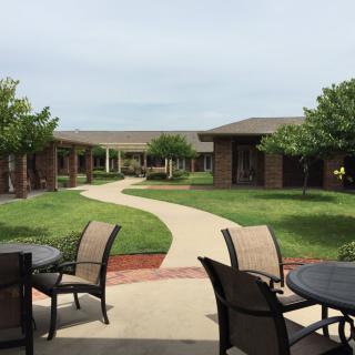 A peaceful courtyard with a winding pathway, surrounded by buildings and greenery, featuring outdoor seating arrangements.