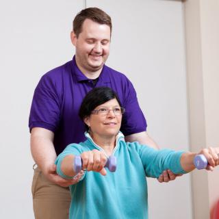 A therapist assists an elderly woman with arm exercises using dumbbells.