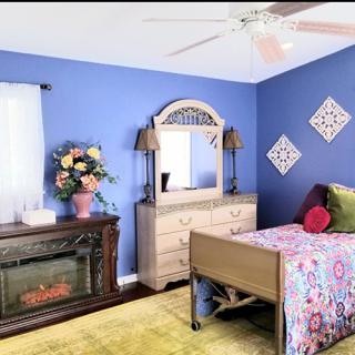 A cozy bedroom with blue walls, featuring a bed with colorful bedding, a dresser with a mirror, and decorative elements like floral arrangements and a fireplace.