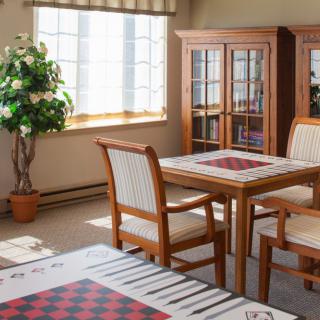 A cozy dining room featuring a chessboard on the table, surrounded by wooden chairs, with natural light streaming through the window.