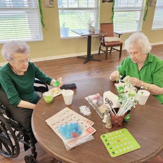 Two elderly women engaged in a crafting activity at a table, surrounded by games and materials.