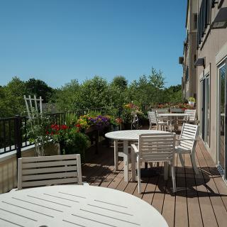 A sunny outdoor patio with white tables and chairs, surrounded by lush greenery.