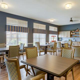 A brightly lit dining room with multiple tables and chairs, featuring large windows and an adjacent kitchen area.