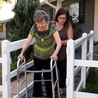 An elderly woman using a walker is assisted down a ramp by a caregiver.