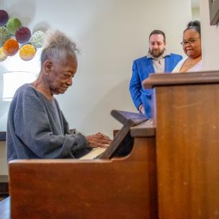 An elderly person playing the piano while two people smile and watch, with colorful balloons in the background.