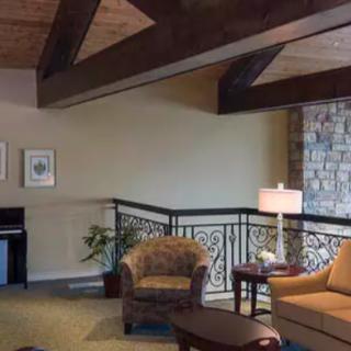 A cozy living room with chairs, a lamp, and a piano, featuring wooden beams and a brick wall.