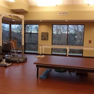 A rehabilitation room equipped with exercise and physical therapy equipment, featuring large windows for natural light.