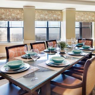 A beautifully set dining table with green dishes and wine glasses, surrounded by upholstered chairs in a bright room with large windows.