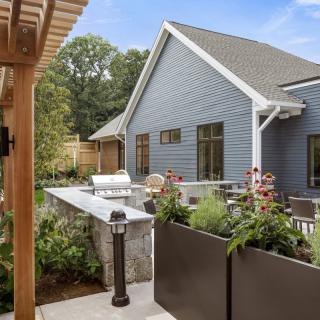 A cozy outdoor patio area featuring a barbecue grill, surrounded by lush plants and a pergola.