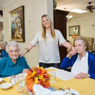 A caregiver stands smiling between two elderly women seated at a dining table, decorated with flowers.