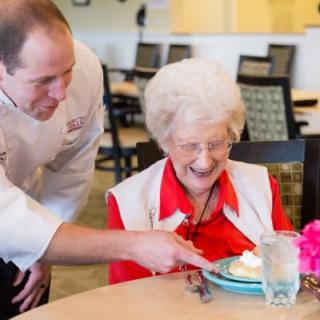 A chef is serving dessert to a smiling elderly woman in a restaurant.
