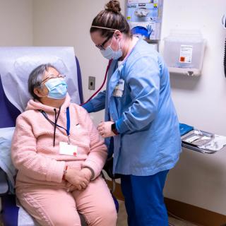 A nurse checks on a patient in a healthcare setting, both wearing masks.