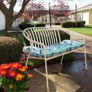 A white metal bench with a floral cushion sits in a sunny garden surrounded by vibrant flowers and trimmed bushes.