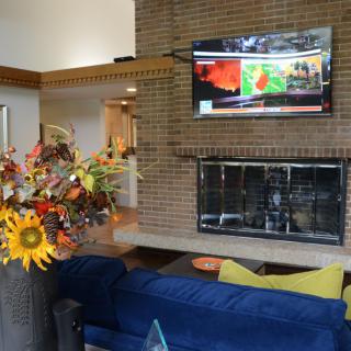 A cozy living room with a brick fireplace and television above, featuring a blue sofa and a vibrant floral arrangement.