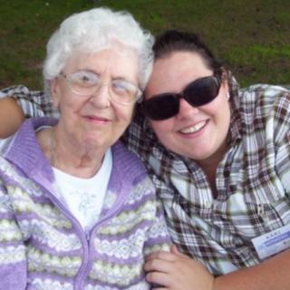 A young woman wearing sunglasses hugs an elderly woman, both smiling outdoors.