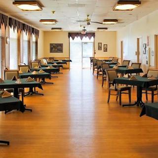 An empty dining hall with neatly arranged tables and chairs, featuring a wooden floor and natural light streaming in through the windows.