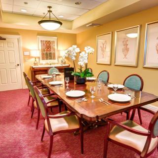 An elegantly decorated dining room with a long wooden table, surrounded by chairs and set with plates. The room features warm lighting, artwork on the walls, and a plush red carpet.