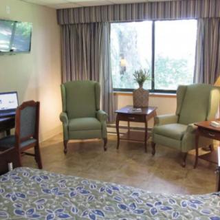 A cozy hotel room featuring a bed, two armchairs, a desk with a television, and a large window.