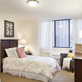 A cozy bedroom with a single bed, lamp, and upholstered chair near a large window.