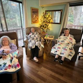 Three seniors enjoying a cozy afternoon by the window, wrapped in colorful quilts.