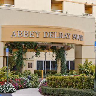 Entrance of a building with 'Abbey Delray South' sign, adorned with plants and flowers.