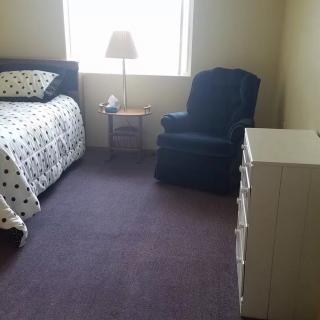 A simple bedroom with a bed, chair, dresser, and lamp by the window.