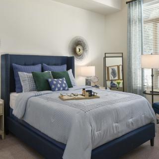 A cozy and well-decorated bedroom featuring a bed with multiple pillows, side lamps, a mirror, and a window with sheer curtains.
