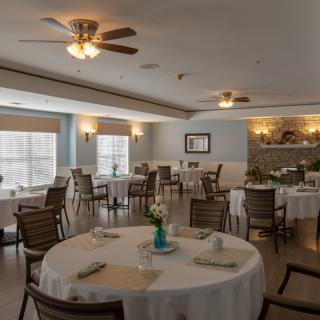 A well-lit dining room with round tables and chairs arranged neatly, featuring ceiling fans and decorative elements.