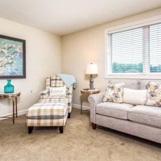 A cozy living room featuring a sofa, armchair with matching ottoman, side tables with decorative items, and a large window.