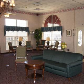 A cozy living room with green sofas and chairs, wooden tables, large windows, and decorative plants.