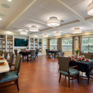 A well-lit dining room with tables, chairs, and a library section featuring bookshelves and a television.
