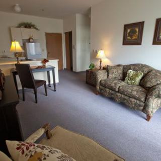 A cozy living room with a patterned couch, lamps, and a small dining area connected to a kitchen.
