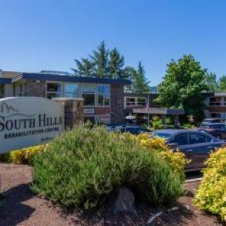 A commercial building with a sign reading 'South Hills,' surrounded by trees, shrubs, and a parking lot with cars.
