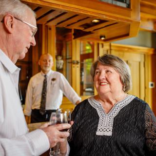 An elderly couple enjoys a conversation over wine in a restaurant, with a bartender in the background.