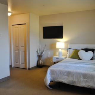 A modern bedroom with a neatly made bed, heart-shaped pillow, and a bedside lamp.