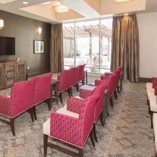 A modern conference room with rows of pink chairs facing a television, featuring large windows with curtains and patterned carpeting.