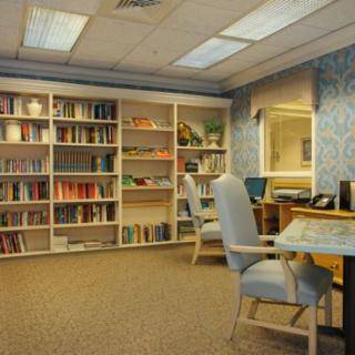 A cozy library room featuring tall bookshelves, a computer desk, and comfortable office chairs under soft fluorescent lighting.