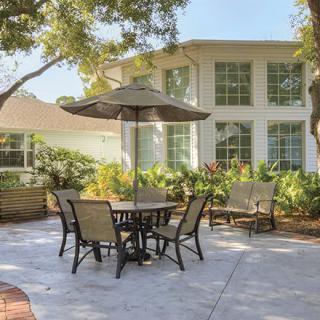 A cozy patio with outdoor furniture under a large umbrella, surrounded by lush greenery and adjacent to a residential building.
