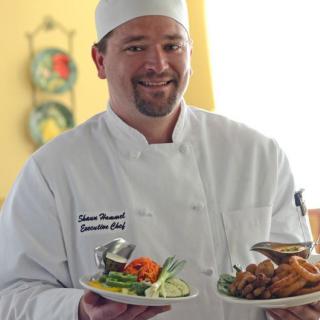 A chef in a white uniform holding two plates of beautifully presented food.
