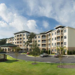 A modern apartment complex with a large parking lot and landscaped surroundings under a blue sky with clouds.