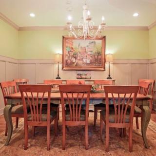 A cozy dining room with a large wooden table, surrounded by matching chairs, illuminated by a chandelier and decorated with artwork on the wall.