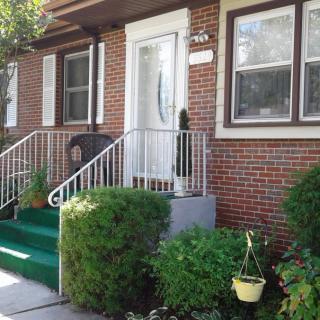 A brick house with a small front porch featuring green carpeted steps, white railings, and various plants.